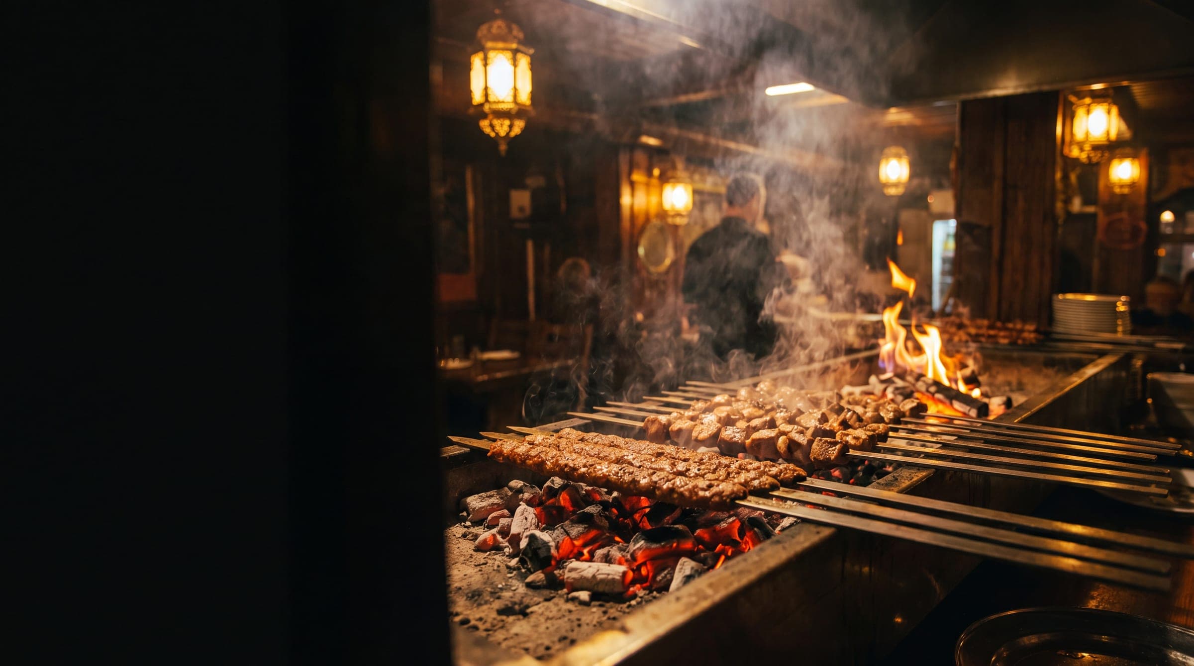 Skewers of adana kebab and lamb shish sizzling over glowing charcoal embers at Origin Mediterranean, with brass lanterns and rising smoke