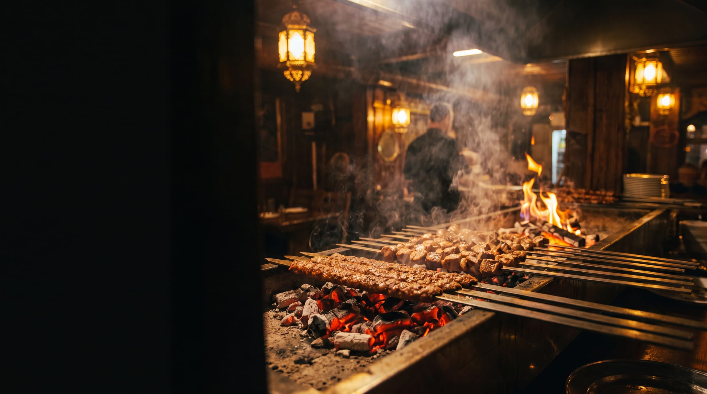 Skewers of adana kebab and lamb shish sizzling over glowing charcoal embers at Origin Mediterranean, with brass lanterns and rising smoke
